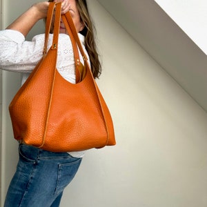Op de afbeelding: Een grote, oranje leren tote bag met een brede, open bovenkant en twee hengsels. De tas is gemaakt van een gestructureerd leer en heeft een casual, ontspannen stijl.