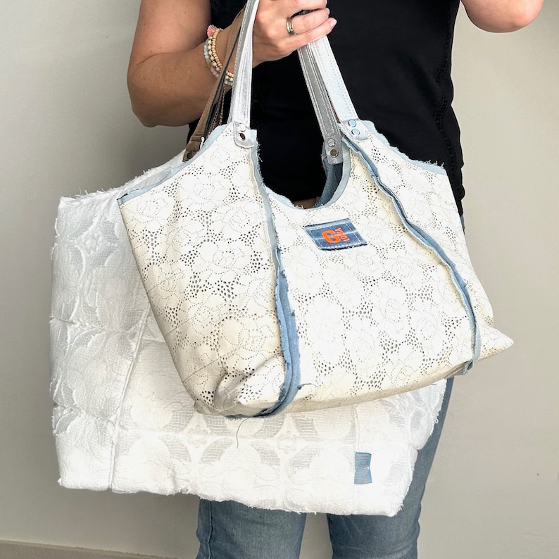 Op de afbeelding: Een witte tote bag met blauwe denim trim en een wit kantpatroon. De tas heeft een klein oranje label met de tekst "G" erop.