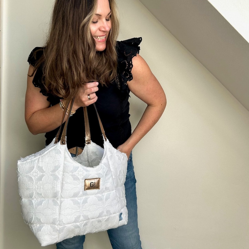 Op de afbeelding: Een witte gewatteerde tote bag met bruine leren handvatten en een gouden monogram "G" aan de voorkant.