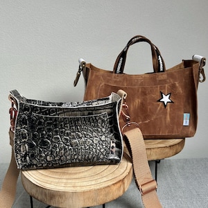 Könnte beinhalten: Zwei Handtaschen auf Holzständern. Eine ist eine braune Tragetasche mit einem weißen Stern und schwarzen Griffen. Die andere ist eine schwarz-graue Umhängetasche mit Krokodilmuster und einem beigen Riemen. Beide Taschen haben Metallbeschläge.
