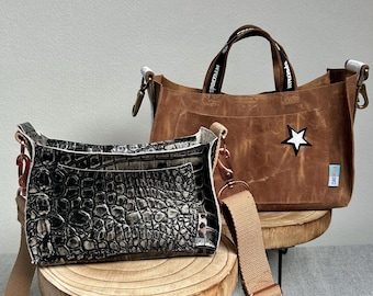 Op de afbeelding: Twee handtassen op houten standaards. Eén is een bruine tote bag met een witte ster en zwarte handvatten. De andere is een zwart-grijze crossbody tas met een krokodillenpatroon en een beige riem. Beide tassen hebben metalen hardware.