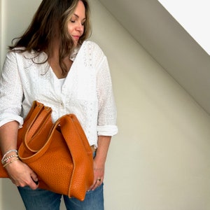 Op de afbeelding: Een vrouw houdt een grote, bruine leren tas met een omslagklep en een bovenste handvat vast. De tas heeft een gestructureerd oppervlak en is gemaakt van hoogwaardig leer.