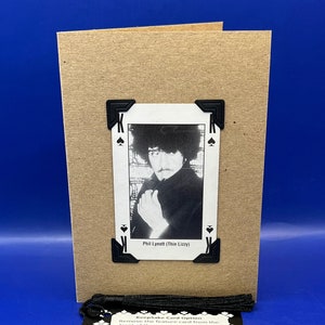May include: A brown card featuring a black and white photograph of Phil Lynott, the lead singer of Thin Lizzy, on the front. The card is decorated with black cardstock corners and a black tassel. The text "Phil Lynott (Thin Lizzy)" is printed below the photo. The back of the card has instructions for turning the card into a bookmark.