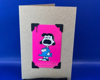 May include: A pink card featuring a black and white illustration of a girl with black hair and a blue dress. The card is decorated with black corners and has the text "19121 UNITED FEATURE SYNDICATE" at the bottom. The card is attached to a brown card with a black tassel.