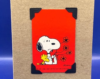 May include: A red card with a black border and corner embellishments features a cartoon image of Snoopy and Woodstock. Snoopy is crying and holding a bouquet of red flowers. The text on the card reads "© 1958, 1965 United Feature Syndicate, Inc."
