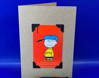 May include: A card with a red background and a cartoon image of a boy wearing a yellow shirt and a blue hat. The card has a black border and the text "MANAGE" is printed on the shirt. The card is attached to a brown card with a black tassel.