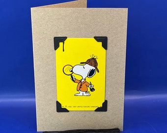 May include: A yellow card with a black and white illustration of Snoopy dressed as a detective. The card has a black tassel attached to it. The text on the card reads "Keepsake Card Option Remove the feature card from the front of the greeting card, make a small hole and use the attached tassel to turn it into a bookmark as per this example. Made by DealtyTheSea"