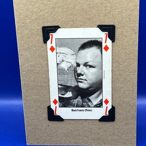 May include: A card featuring a black and white photograph of Black Francis from the Pixies. The card is a 7 of diamonds and is secured with black corner protectors.