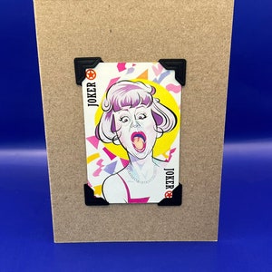 May include: A brown card with a joker card image on it. The joker card features a woman with pink hair and a bright yellow background. The card is decorated with colourful geometric shapes. The text "JOKER" is printed on the card in black.