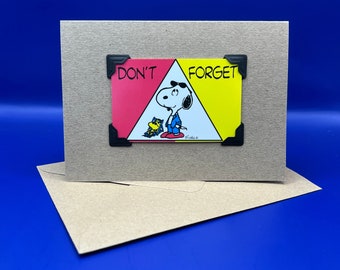 May include: A card featuring a cartoon image of Snoopy and Woodstock. The card is divided into two triangles, one red and one yellow. The red triangle says "Don't" and the yellow triangle says "Forget".