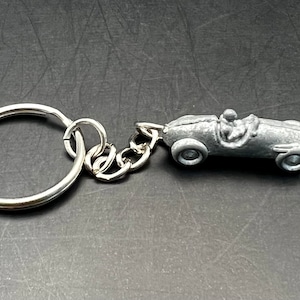 May include: A silver-coloured keyring featuring a miniature race car with a driver. The car has visible wheels and is attached to a chain and a large key ring. The keyring is made of metal.