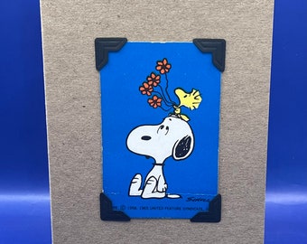 May include: A card featuring a vintage cartoon image of Snoopy holding a bunch of red flowers on his head. The image is framed with black corners and includes the text "© 1958, 1965 UNITED FEATURE SYNDICATE, INC."