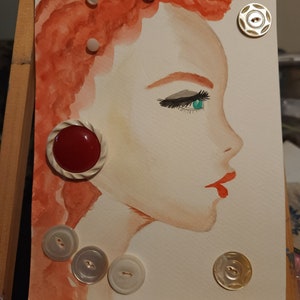 May include: Watercolour painting of a woman's face with red hair and green eyes. The painting is decorated with various buttons, including red, white and gold.