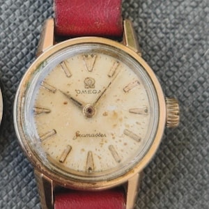 Könnte beinhalten: Vintage Omega Seamaster Uhr in Gold mit cremefarbenem Zifferblatt und rotem Lederarmband. Das Zifferblatt hat goldene Markierungen und Zeiger, das Omega-Logo und den Schriftzug "Seamaster". Eine klassische Armbanduhr.