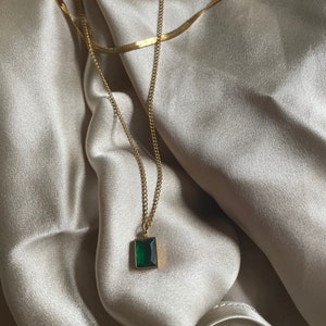 Op de afbeelding: Een gouden ketting met een groene edelsteen hanger. De ketting ligt op een beige stoffen achtergrond.