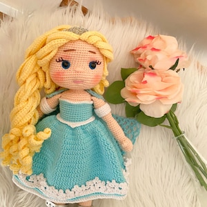 Puede incluir: Mu&ntilde;eca de ganchillo hecha a mano con pelo largo amarillo, ojos azules y un vestido azul claro con ribete blanco. La mu&ntilde;eca lleva zapatos plateados y una tiara plateada. Un ramo de rosas rosas est&aacute; cerca.