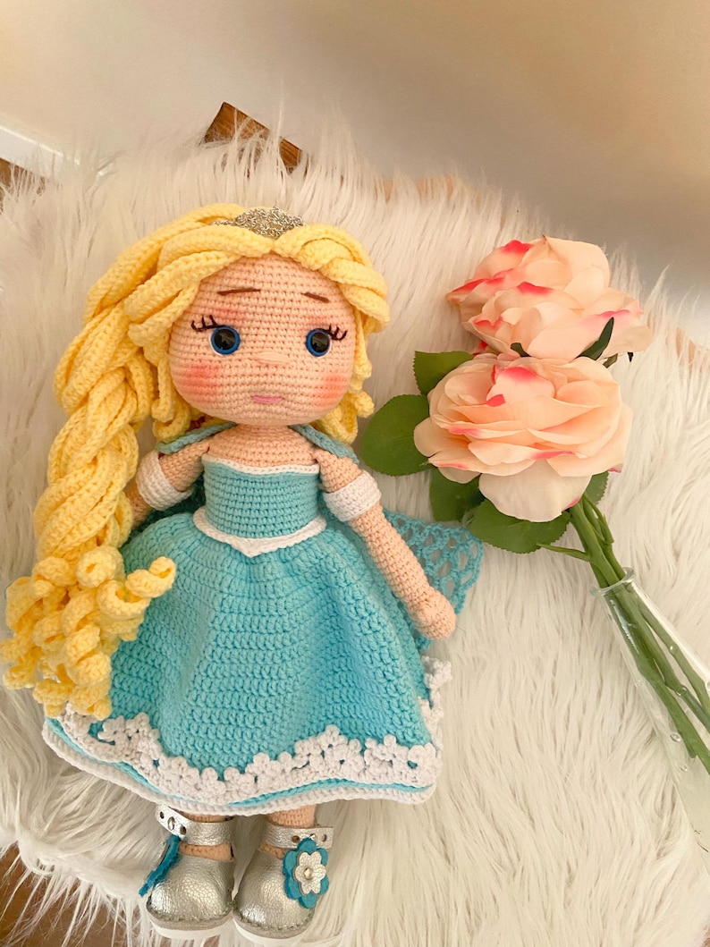 Puede incluir: Mu&ntilde;eca de ganchillo hecha a mano con pelo largo amarillo, ojos azules y un vestido azul claro con ribete blanco. La mu&ntilde;eca lleva zapatos plateados y una tiara plateada. Un ramo de rosas rosas est&aacute; cerca.