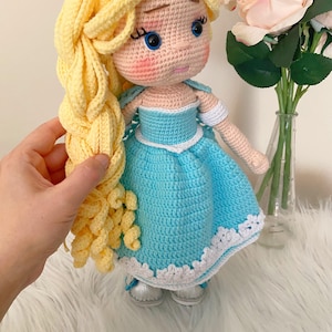 Puede incluir: Mu&ntilde;eca hecha a mano con ganchillo, con pelo largo y rubio y un vestido azul claro con ribete blanco. La mu&ntilde;eca tiene una tiara plateada, ojos azules y zapatos plateados. Juguete con tem&aacute;tica de princesa.