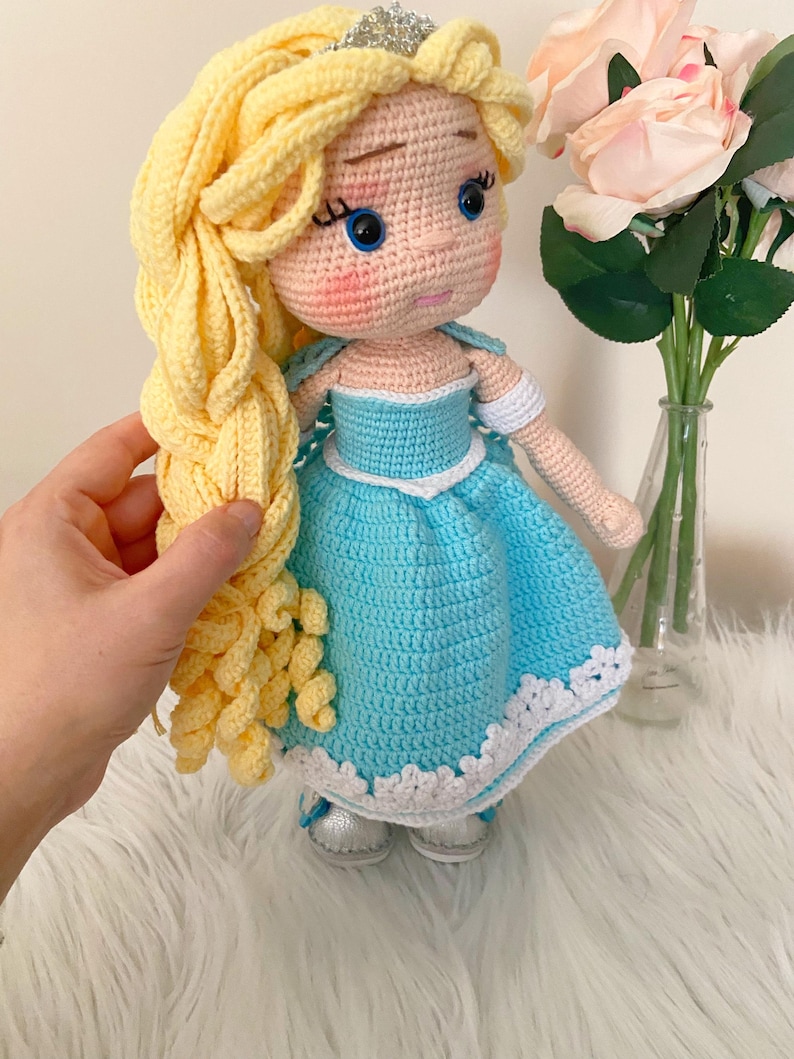 Puede incluir: Mu&ntilde;eca hecha a mano con ganchillo, con pelo largo y rubio y un vestido azul claro con ribete blanco. La mu&ntilde;eca tiene una tiara plateada, ojos azules y zapatos plateados. Juguete con tem&aacute;tica de princesa.