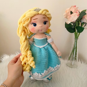 Puede incluir: Mu&ntilde;eca de ganchillo hecha a mano con cabello rubio peinado en una trenza. La mu&ntilde;eca lleva un vestido azul claro con ribete blanco y zapatos plateados. La mu&ntilde;eca tiene ojos azules y una corona plateada. La mu&ntilde;eca mide unos 30 cm.