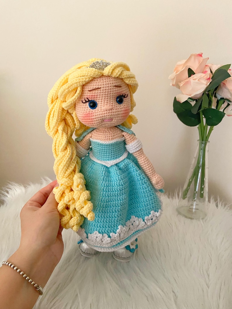 Puede incluir: Mu&ntilde;eca de ganchillo hecha a mano con cabello rubio peinado en una trenza. La mu&ntilde;eca lleva un vestido azul claro con ribete blanco y zapatos plateados. La mu&ntilde;eca tiene ojos azules y una corona plateada. La mu&ntilde;eca mide unos 30 cm.