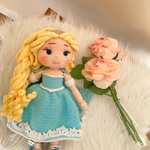Puede incluir: Mu&ntilde;eca de ganchillo hecha a mano con pelo largo y amarillo y una tiara plateada. La mu&ntilde;eca lleva un vestido azul claro con ribete de encaje blanco y zapatos plateados. Un ramo de rosas rosas en un jarr&oacute;n de cristal est&aacute; cerca.