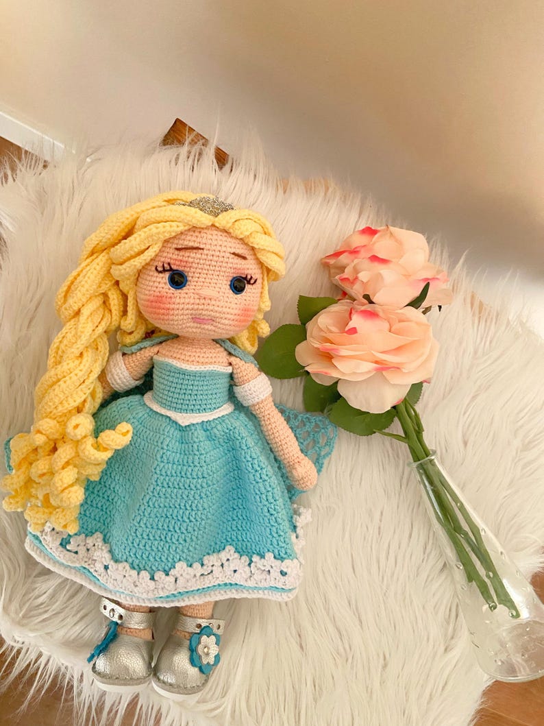 Puede incluir: Mu&ntilde;eca de ganchillo hecha a mano con pelo largo y amarillo y una tiara plateada. La mu&ntilde;eca lleva un vestido azul claro con ribete de encaje blanco y zapatos plateados. Un ramo de rosas rosas en un jarr&oacute;n de cristal est&aacute; cerca.