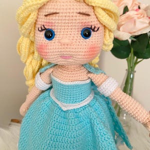 Puede incluir: Mu&ntilde;eca de ganchillo hecha a mano con piel clara, ojos azules y cabello rubio peinado en una trenza. La mu&ntilde;eca viste un vestido azul claro con ribetes blancos y una tiara plateada. Las mejillas de la mu&ntilde;eca son rosadas.