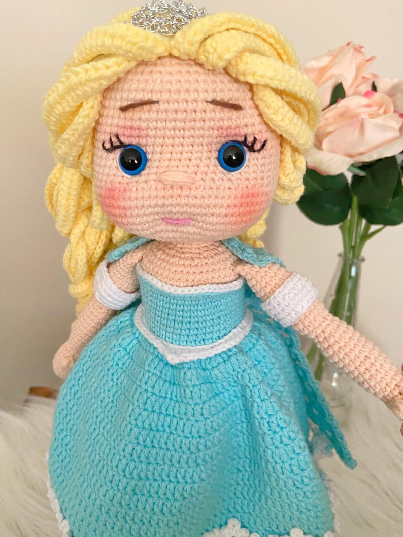 Puede incluir: Mu&ntilde;eca de ganchillo hecha a mano con piel clara, ojos azules y cabello rubio peinado en una trenza. La mu&ntilde;eca viste un vestido azul claro con ribetes blancos y una tiara plateada. Las mejillas de la mu&ntilde;eca son rosadas.
