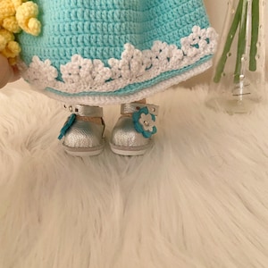 Puede incluir: Mu&ntilde;eca hecha a mano con un vestido de ganchillo azul claro, ribete de encaje blanco y zapatos plateados. Los zapatos tienen un peque&ntilde;o adorno de flor azul. El pelo de la mu&ntilde;eca es amarillo.