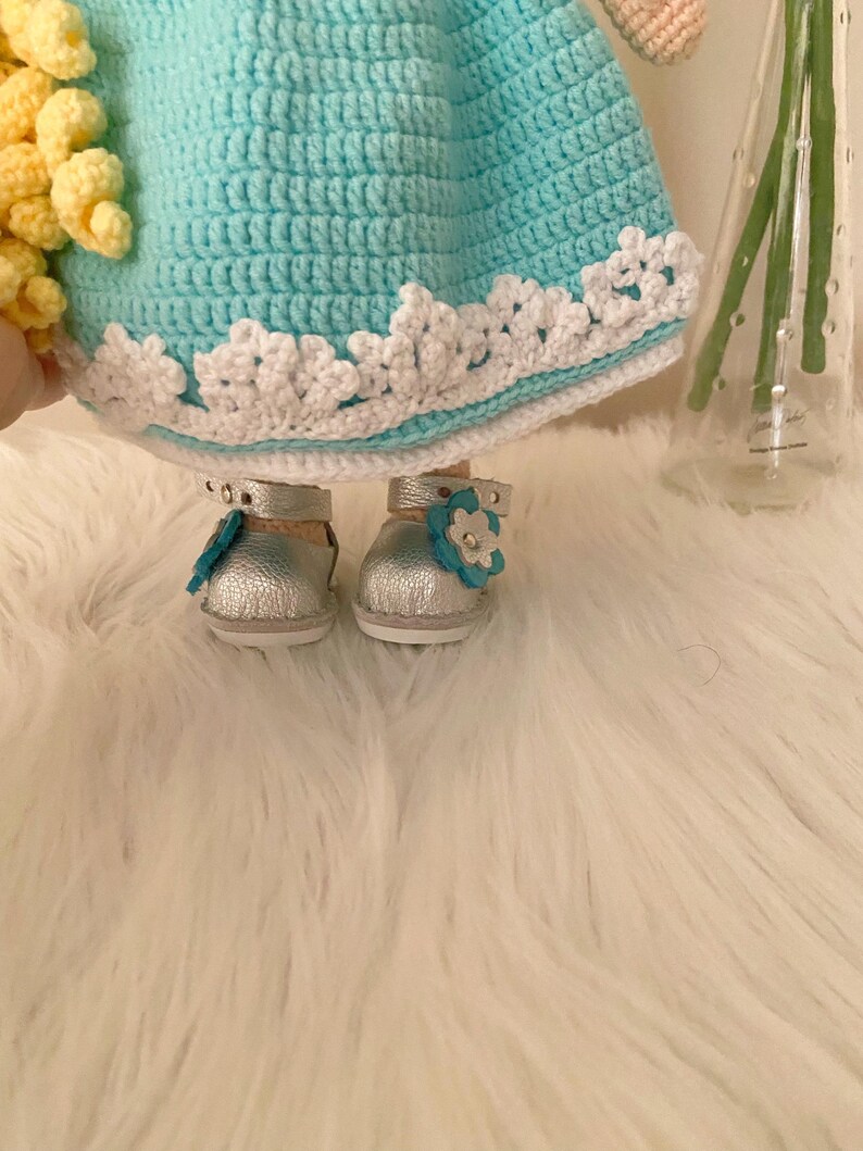 Puede incluir: Mu&ntilde;eca hecha a mano con un vestido de ganchillo azul claro, ribete de encaje blanco y zapatos plateados. Los zapatos tienen un peque&ntilde;o adorno de flor azul. El pelo de la mu&ntilde;eca es amarillo.