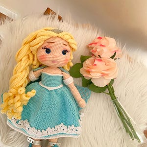 Puede incluir: Una mu&ntilde;eca de ganchillo hecha a mano con pelo largo y amarillo y un vestido azul. La mu&ntilde;eca tiene una tiara plateada y zapatos plateados. Un ramo de rosas rosas en un jarr&oacute;n de cristal transparente est&aacute; junto a la mu&ntilde;eca.