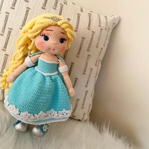 Puede incluir: Mu&ntilde;eca de ganchillo hecha a mano con cabello rubio, ojos azules y un vestido azul claro con ribete blanco. La mu&ntilde;eca tiene zapatos plateados y una tiara dorada. La mu&ntilde;eca est&aacute; sentada sobre una superficie blanca y esponjosa.