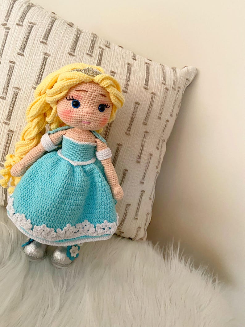 Puede incluir: Mu&ntilde;eca de ganchillo hecha a mano con cabello rubio, ojos azules y un vestido azul claro con ribete blanco. La mu&ntilde;eca tiene zapatos plateados y una tiara dorada. La mu&ntilde;eca est&aacute; sentada sobre una superficie blanca y esponjosa.
