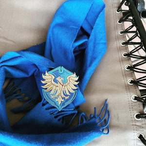 May include: A bright blue scarf with fringed ends, featuring a decorative patch. The patch displays a gold eagle emblem on a shield-shaped background with blue and teal details. The scarf is laid on a neutral-coloured surface.