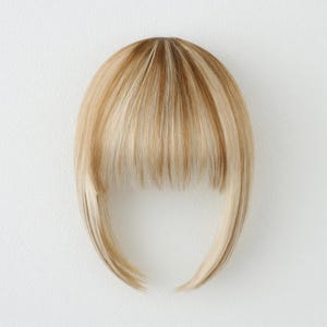 May include: A blonde hairpiece with a layered fringe and long side pieces. The hair has a mix of light and medium blonde tones. The hairpiece is designed to be worn on the forehead, offering a natural look.