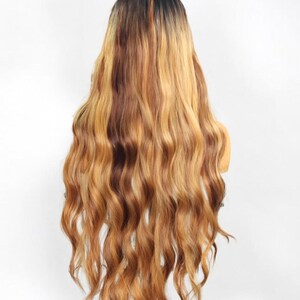 May include: A long, wavy wig featuring a dark brown root color that transitions into a multi-tonal blend of golden blonde and light brown highlights. The hair has a soft, flowing texture with loose waves extending down to the lower back area.