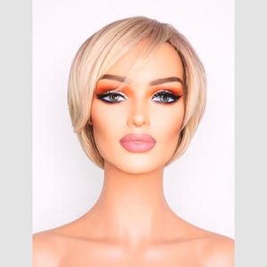 Könnte beinhalten: Ein Schaufensterpuppenkopf mit einer kurzen, gestuften blonden Perücke. Die Perücke hat einen Seitenscheitel und ist mit weichen Wellen gestylt. Die Schaufensterpuppe hat ein komplettes Make-up, einschließlich orangefarbenem und goldenem Lidschatten, schwarzem Eyeliner und rosa Lippenstift. Der Hintergrund ist weiß.