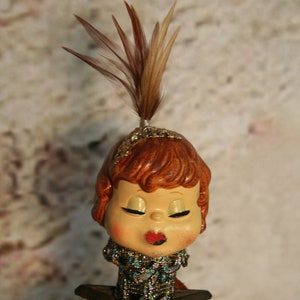 May include: A ceramic doll head with brown hair and a feather headdress. The doll is wearing a beaded necklace and has a red painted mouth. The doll is holding a box of sage with the text "Sage Pure Ground" printed on it.