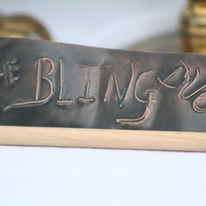 May include: A metal sign with the words "The Bling Duo" etched into the surface. The sign is made of a dark metal and has a rustic look.