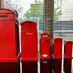 May include: A set of six red miniature replica British telephone boxes, ranging in size from large to small. The boxes are made of metal and have a traditional design.