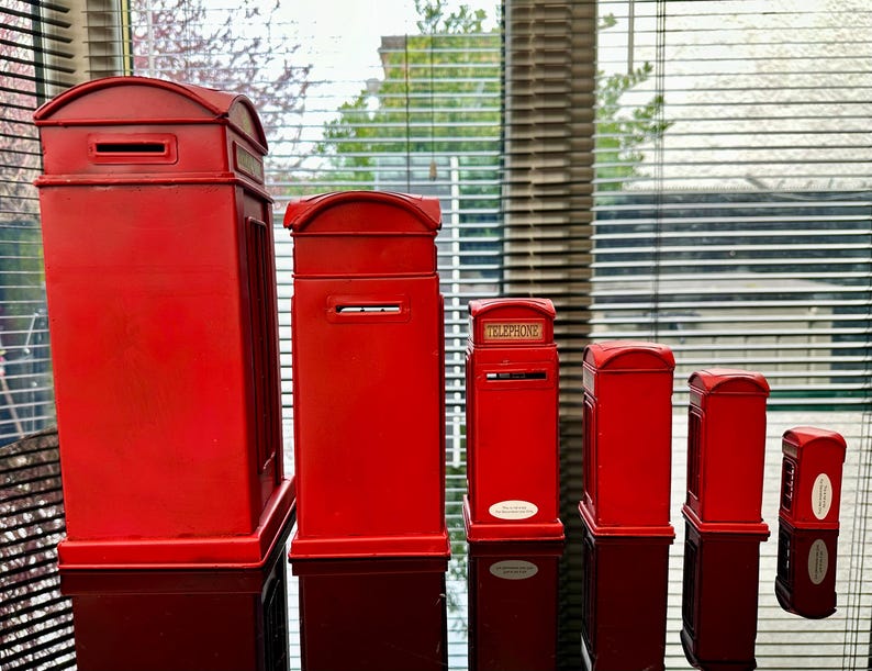 May include: A set of six red miniature replica British telephone boxes, ranging in size from large to small. The boxes are made of metal and have a traditional design.