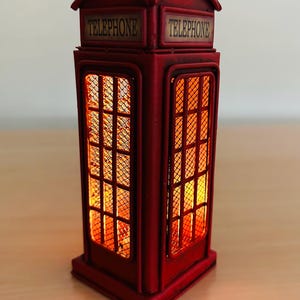Könnte beinhalten: Eine rote Metall-Replik einer klassischen britischen Telefonzelle mit einem Licht im Inneren. Die Telefonzelle hat auf beiden Seiten den Schriftzug "TELEPHONE" aufgedruckt.
