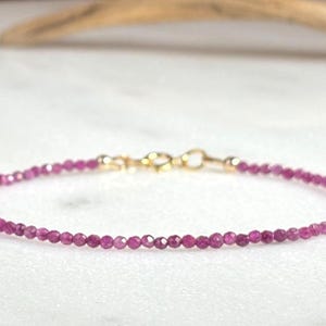 Peut inclure: Bracelet délicat orné de petites pierres précieuses rondes rose foncé. Le bracelet est doté d'un fermoir doré. Le bijou est présenté sur une surface blanche.