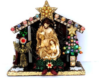 May include: A Christmas nativity scene figurine featuring the Holy Family, an angel, and a Christmas tree. The figurine is decorated with colorful jewels and a gold star. The base is gold-colored with a textured finish.