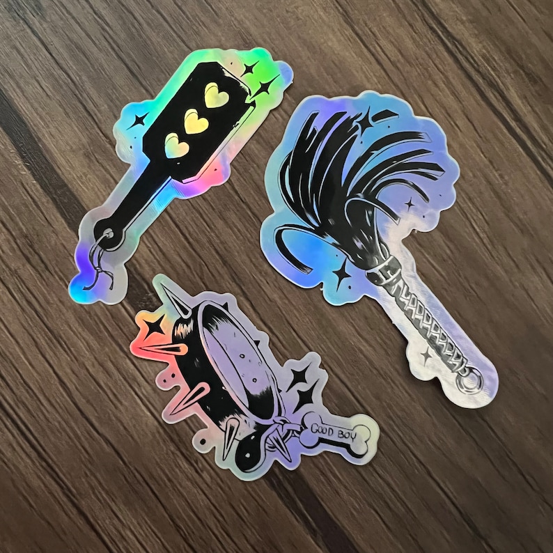 May include: Three iridescent stickers with black and white illustrations. The first sticker shows a paddle with three hearts on it. The second sticker shows a whip with a braided handle. The third sticker shows a collar with spikes and the text "Good Boy".