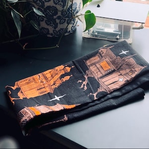 May include: Black fabric kitchen towel with an orange and white print of houses and stars. The print is reminiscent of a spooky Halloween theme.