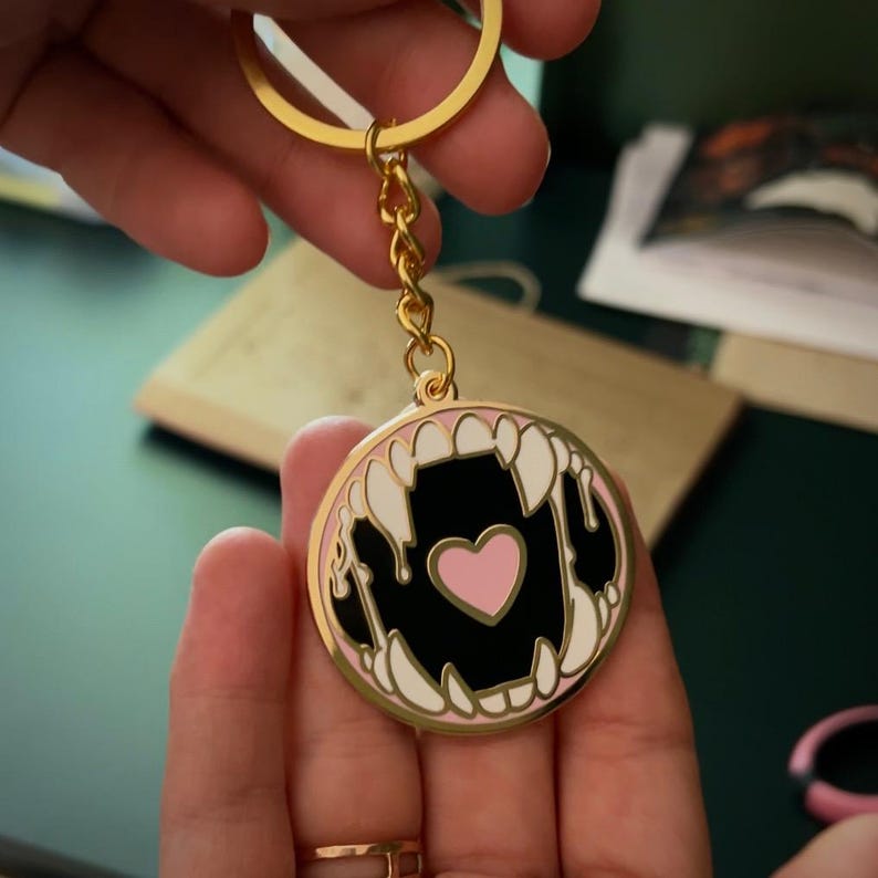 May include: Gold keychain with a round enamel charm featuring a black and white image of a mouth with fangs and a pink heart inside.