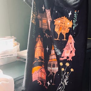 May include: Black tea towel with a whimsical design of orange and pink houses with legs on a black background. The houses have white accents and are surrounded by white stars and green foliage.