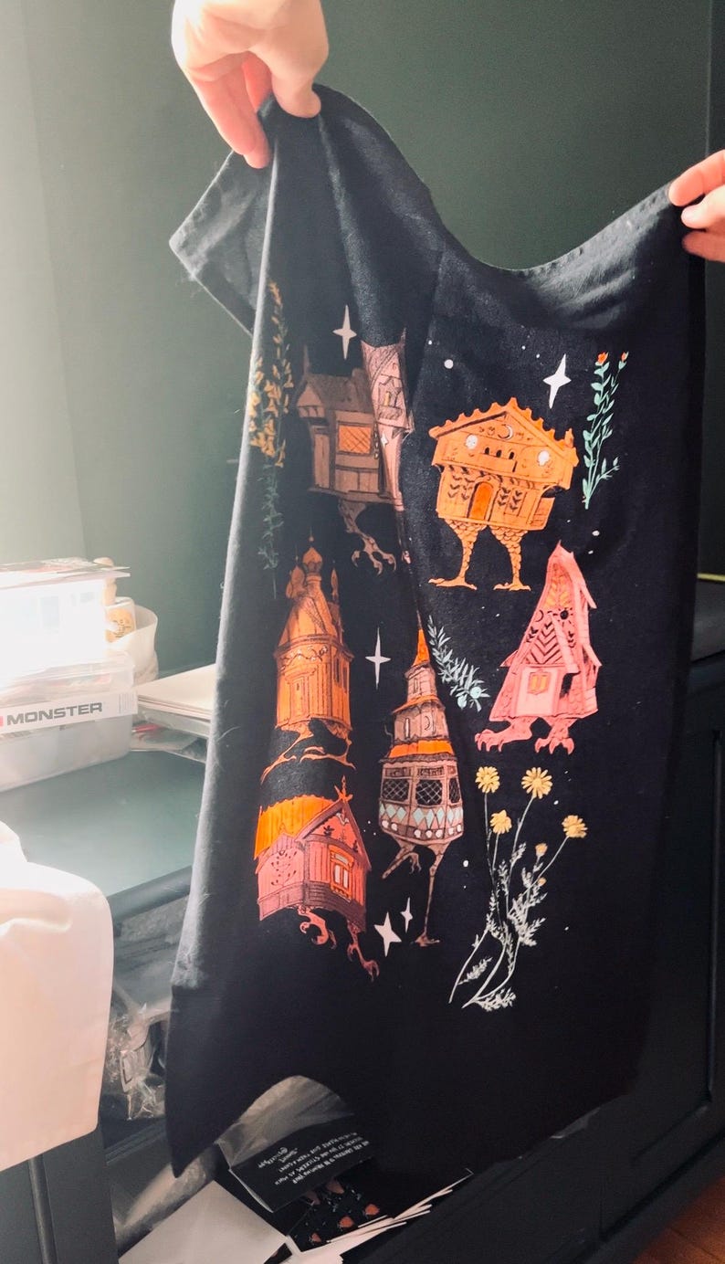 May include: Black tea towel with a whimsical design of orange and pink houses with legs on a black background. The houses have white accents and are surrounded by white stars and green foliage.